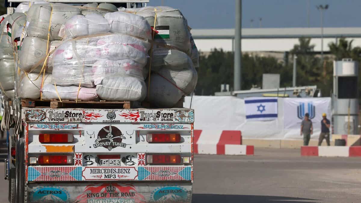 Aid truck loaded with supplies approaches Israeli checkpoint, with flags and guards in the background.