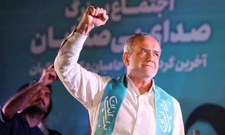 Photo of Iran Masoud Pezeshkian raising his fist during a campaign rally in Iran, wearing a turquoise sash with Persian writing, standing in front of a teal backdrop with Persian text.