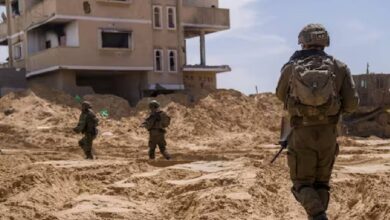 Israeli soldiers patrol a sandy, war-torn area in Rafah, Gaza Strip, near a damaged residential building during a military operation under a clear blue sky.