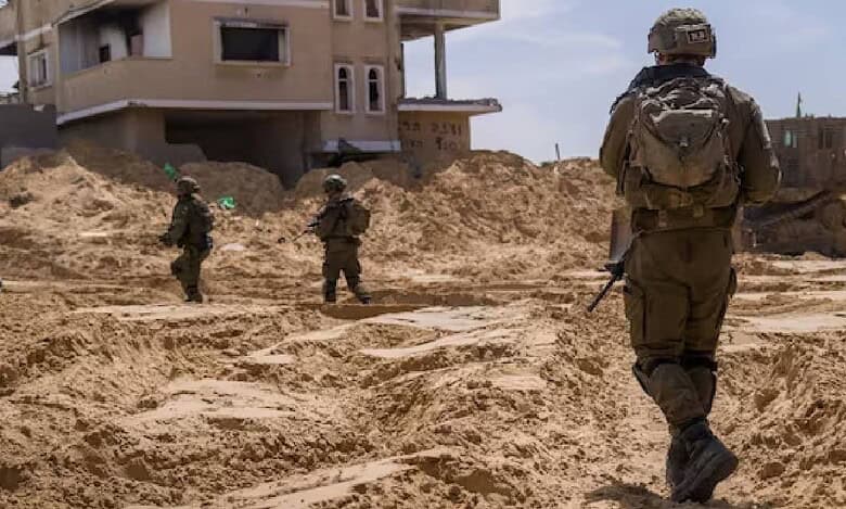 Israeli soldiers patrol a sandy, war-torn area in Rafah, Gaza Strip, near a damaged residential building during a military operation under a clear blue sky.