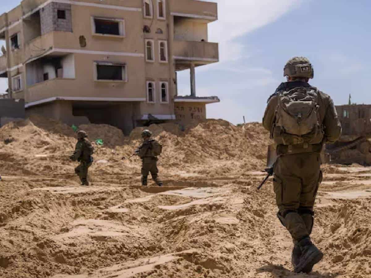 Israeli soldiers patrol a sandy, war-torn area in Rafah, Gaza Strip, near a damaged residential building during a military operation under a clear blue sky.