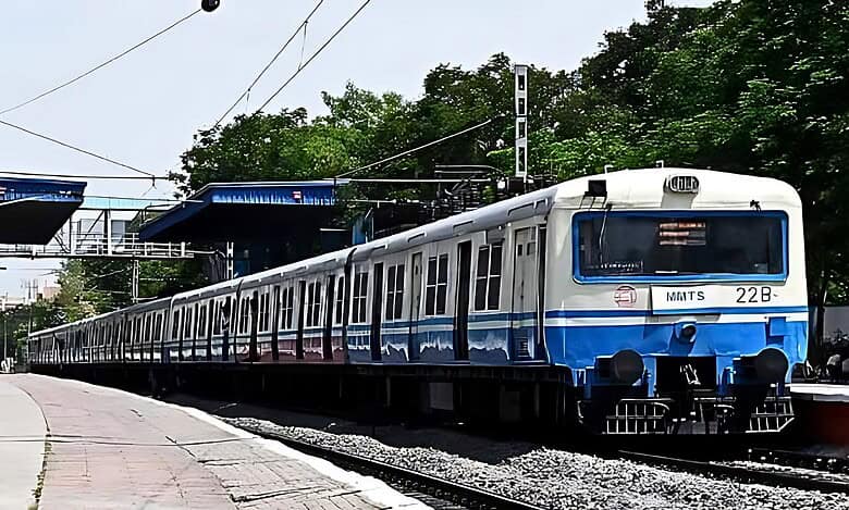 MMTS train parked at a Hyderabad platform with blue and white livery, under a canopy with green trees behind.