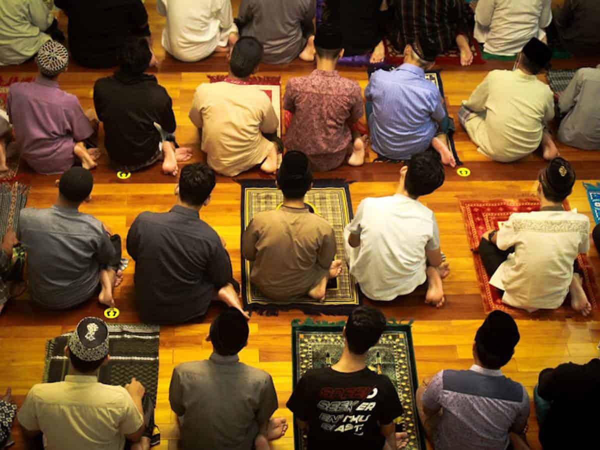 Group of men sitting on prayer mats in a mosque, preparing for congregational prayer. The image shows rows of worshippers from a top angle, wearing traditional and casual attire on a polished wooden floor.