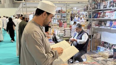 Visitor reading a book at a stall during the 29th Muscat International Book Fair, with shelves of Arabic books and exhibitors in the background.