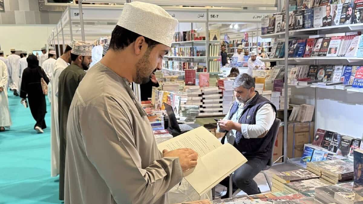 Visitor reading a book at a stall during the 29th Muscat International Book Fair, with shelves of Arabic books and exhibitors in the background.
