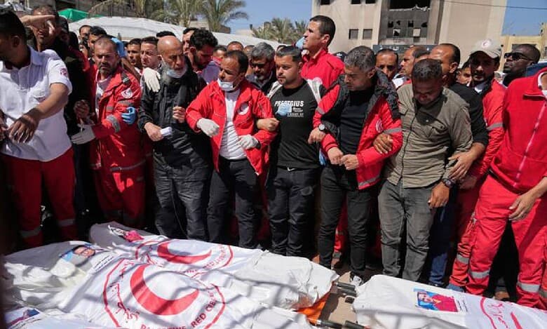 A group of people, some in medical uniforms, stand in solidarity behind several bodies covered in Palestine Red Crescent shrouds during a public funeral.