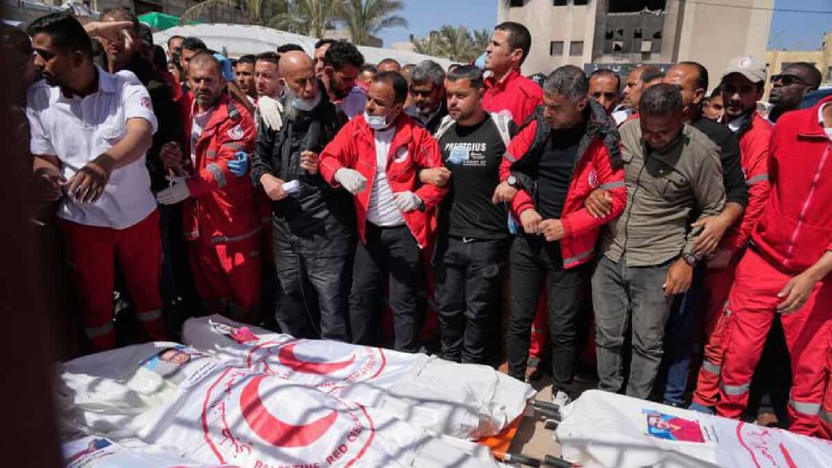 A group of people, some in medical uniforms, stand in solidarity behind several bodies covered in Palestine Red Crescent shrouds during a public funeral.