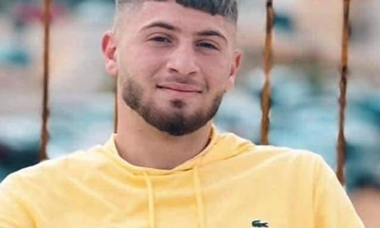 Portrait of a Palestinian teen with short hair and trimmed beard, wearing a yellow Lacoste hoodie, standing in front of a rusted fence with an urban background.