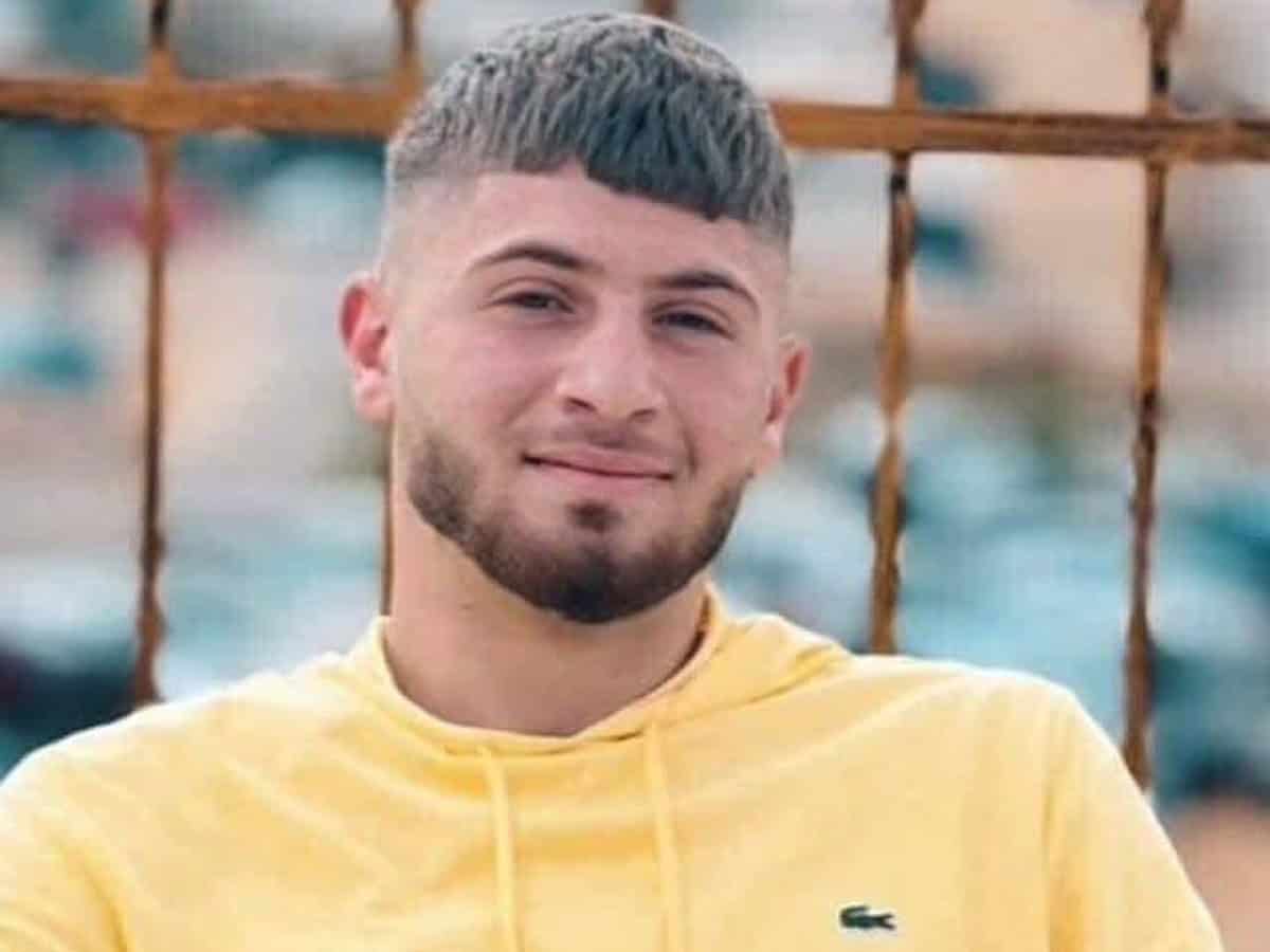 Portrait of a Palestinian teen with short hair and trimmed beard, wearing a yellow Lacoste hoodie, standing in front of a rusted fence with an urban background.