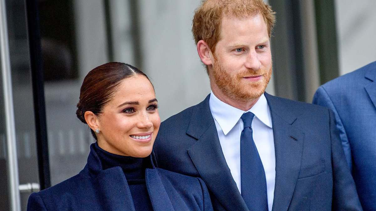 Photo of Prince Harry and Meghan Markle wearing formal navy outfits, smiling and standing together outside a building.