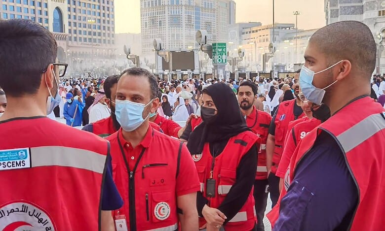 Saudi Red Crescent Authority volunteers in red vests and face masks coordinating efforts during Haj season in Makkah, Saudi Arabia, with pilgrims in the background.