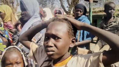 Displaced Sudanese children and women look distressed while standing outdoors during the ongoing conflict.