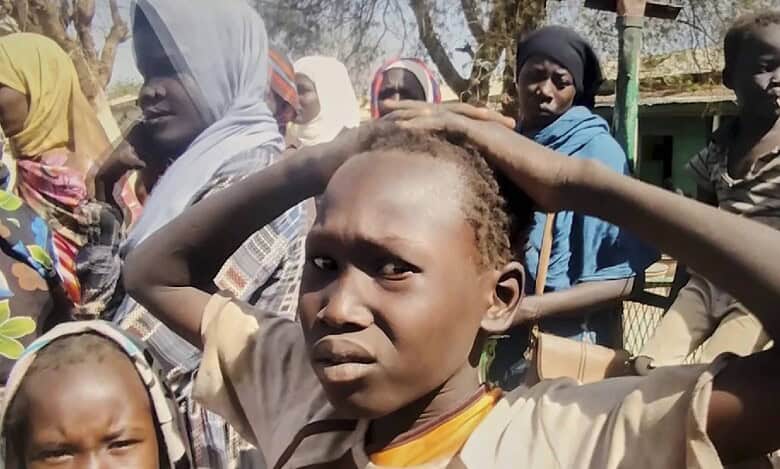 Displaced Sudanese children and women look distressed while standing outdoors during the ongoing conflict.