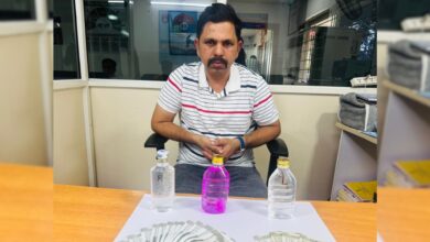 A man in a striped shirt sits at a desk with Rs 2,22,000 in cash, two water bottles labeled "L" and "R," and a pink liquid bottle, linked to a bribery arrest by Shamirpet police.
