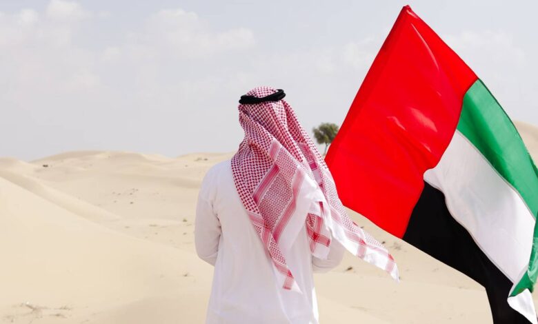 A man wearing traditional Emirati attire walks in the desert carrying the UAE national flag, under a partly cloudy sky.