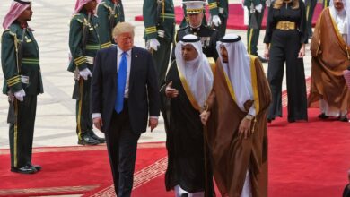 Donald Trump walks alongside Saudi King Salman on a red carpet at King Khalid International Airport, flanked by officials and ceremonial guards, with Melania Trump following behind.