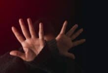 Person shielding their face with open hands in a defensive gesture, lit by red light against a dark background.