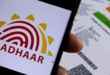 Close-up of a smartphone showing the Aadhaar logo, with printed Aadhaar cards in the background on a wooden surface.