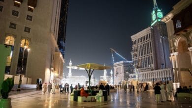 Night view of pilgrims using the new walkway from Address Jabal Omar Makkah to the Grand Mosque, with the iconic clock tower illuminated in the background.