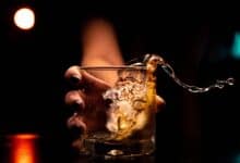 A close-up of a hand holding a glass of alcoholic drink with ice, as the liquid splashes out dramatically against a dark background.