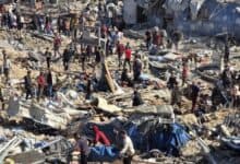 A chaotic scene of destruction in Gaza, with dozens of Palestinians standing amidst rubble and debris from recent Israeli airstrikes. Collapsed buildings, scattered belongings, and makeshift shelters are visible, as people appear to search for survivors or belongings.
