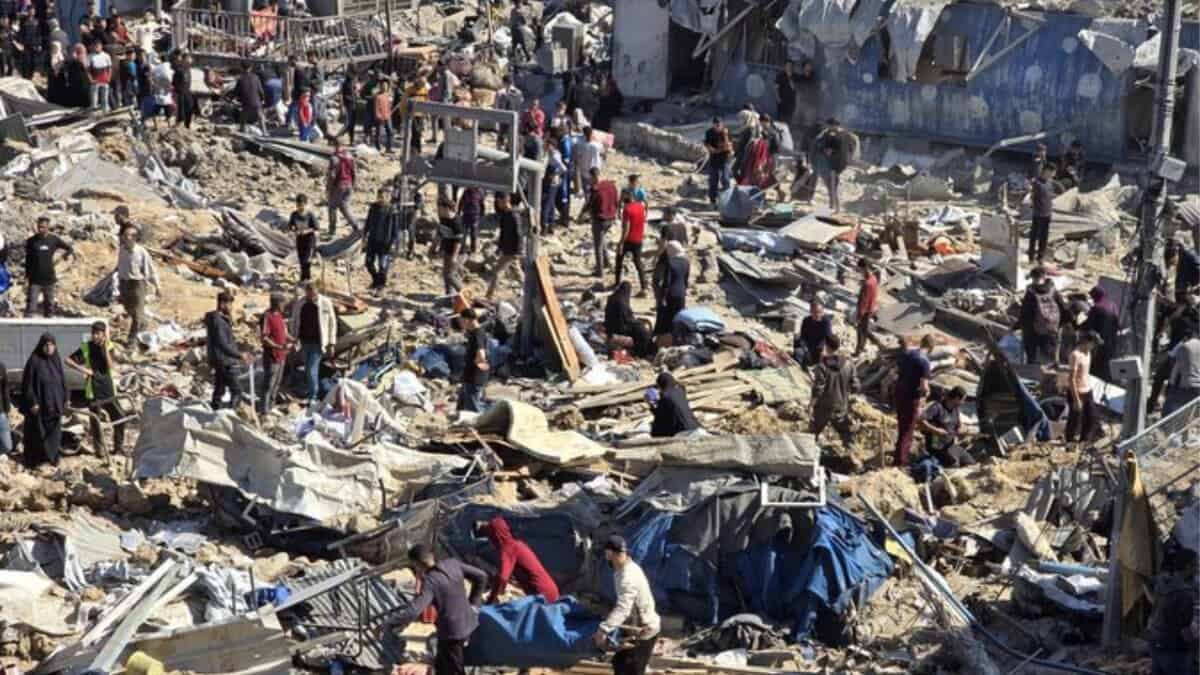 A chaotic scene of destruction in Gaza, with dozens of Palestinians standing amidst rubble and debris from recent Israeli airstrikes. Collapsed buildings, scattered belongings, and makeshift shelters are visible, as people appear to search for survivors or belongings.