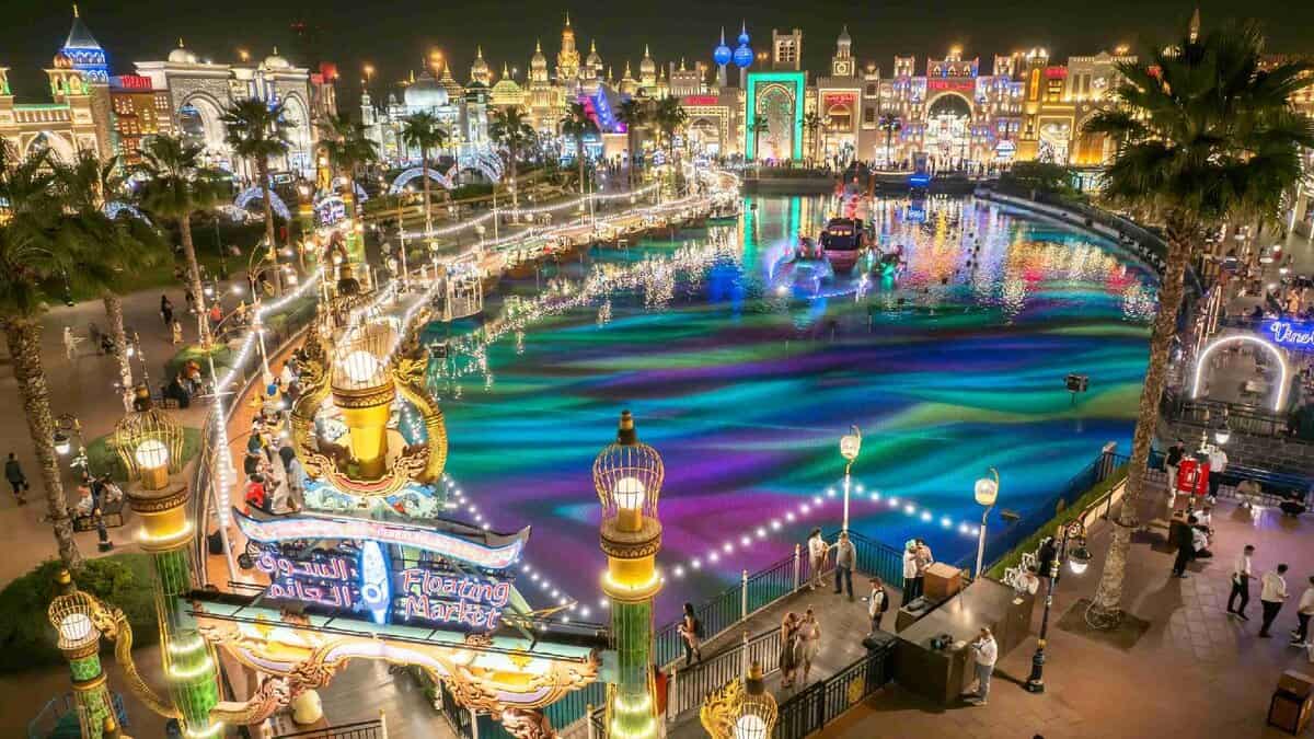 A vibrant night view of Dubai's Global Village featuring the Floating Market, colourful water show, and illuminated pavilions representing various countries.