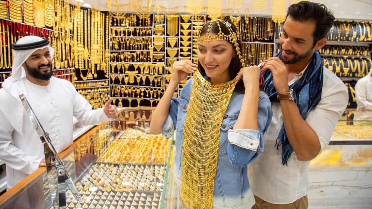 A woman tries on a gold necklace and headpiece in a Dubai jewellery shop, helped by a man, as a smiling shopkeeper gives a thumbs-up.