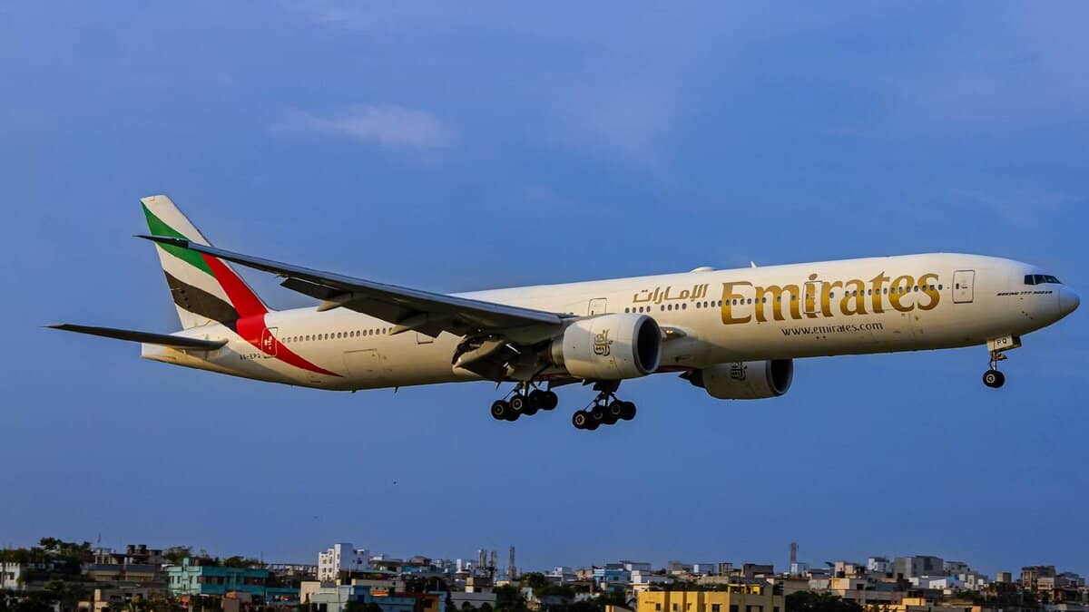 Emirates Boeing 777 aircraft flying low over a cityscape during landing, with landing gear deployed.