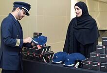 An Emirates pilot in uniform receives a commemorative cap from a staff member during World Pilots' Day celebrations, with branded ceramic mugs and caps displayed on a table.