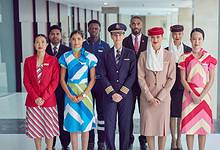 A diverse group of Emirates Group staff, including pilots, cabin crew, and ground personnel, standing in uniform at a modern airport terminal. The image showcases multicultural representation and professional airline attire.