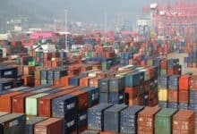 Hundreds of colorful shipping containers stacked at a busy port terminal with cranes and a hazy cityscape in the background