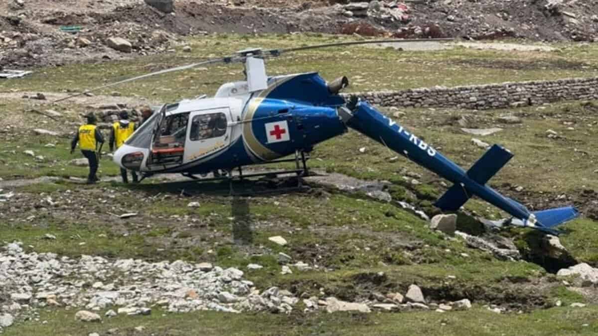 A medical helicopter with a red cross, crashed on a grassy, rocky terrain in Kedarnath. Two rescuers in yellow vests assist near the wreckage.