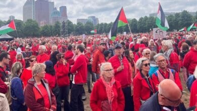 Large crowd dressed in red gathers at a protest with Palestinian flags and signs calling for an end to genocide, under cloudy skies.