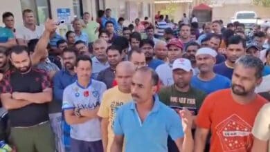 A large group of Indian migrant workers standing outside a building in Saudi Arabia. One man in a blue shirt is speaking while others listen intently.