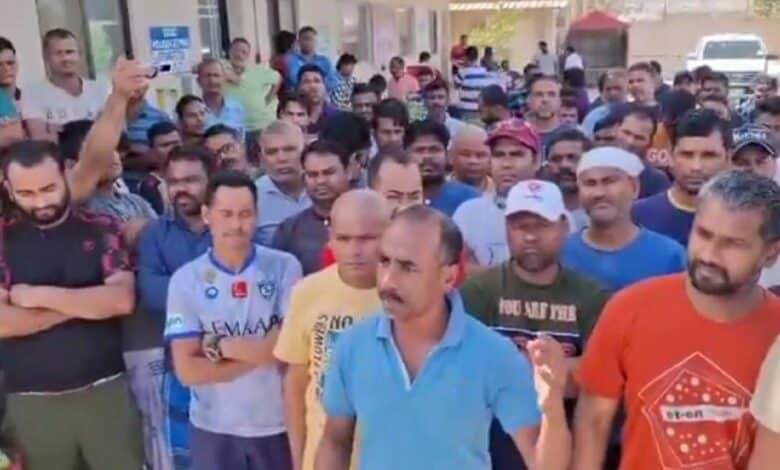 A large group of Indian migrant workers standing outside a building in Saudi Arabia. One man in a blue shirt is speaking while others listen intently.