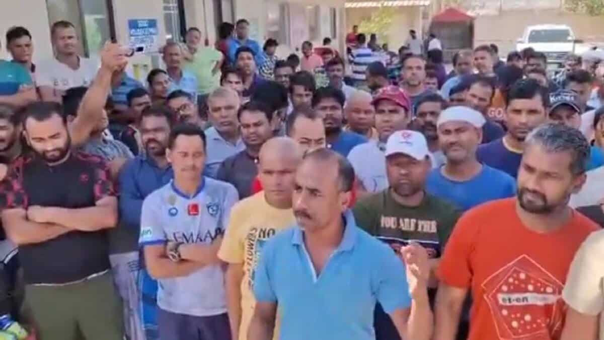 A large group of Indian migrant workers standing outside a building in Saudi Arabia. One man in a blue shirt is speaking while others listen intently.