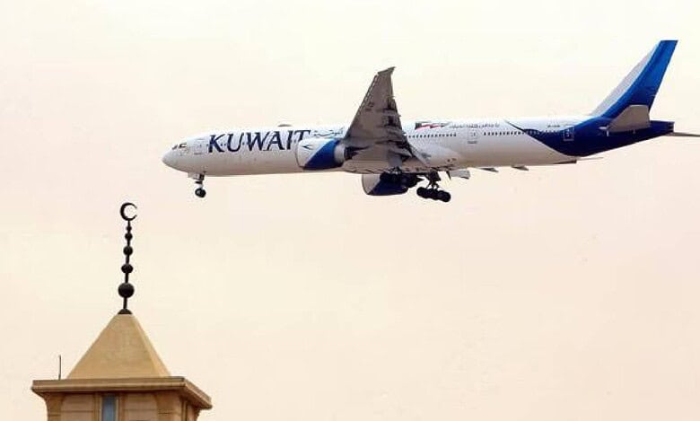 Kuwait Airways aircraft flying over mosque minaret with crescent moon symbol, approaching landing during daylight.