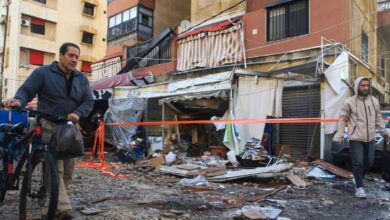 Damaged shopfronts and debris in Beirut, Lebanon, after suspected conflict-related incident, with civilians walking past rubble and cordoned-off area