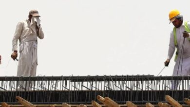 Two migrant workers in Saudi Arabia labour on a construction site under harsh sun, one wiping sweat and the other using equipment.