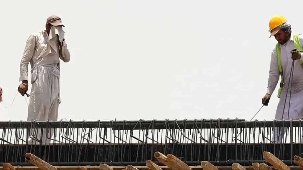 Two migrant workers in Saudi Arabia labour on a construction site under harsh sun, one wiping sweat and the other using equipment.