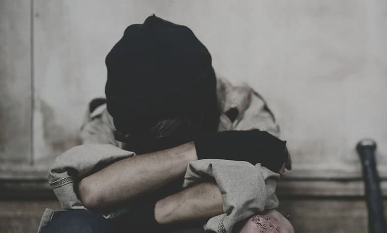 A distressed person sits curled up on the floor with head buried in arms, wearing a black beanie and torn clothing, suggesting emotional trauma or abuse.