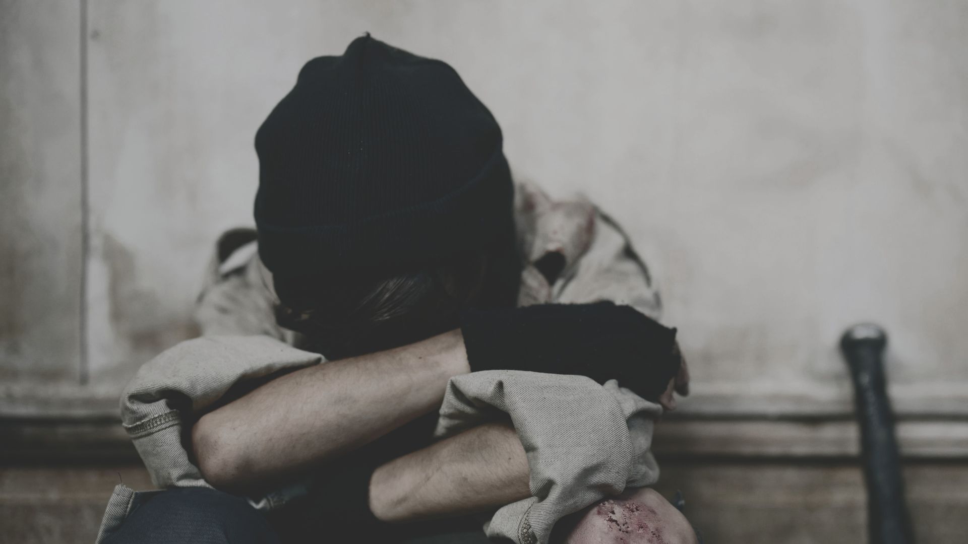 A distressed person sits curled up on the floor with head buried in arms, wearing a black beanie and torn clothing, suggesting emotional trauma or abuse.