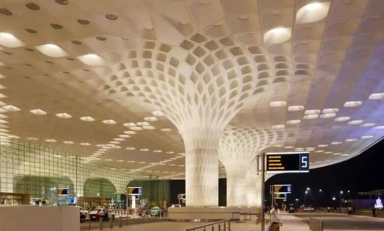 A spacious airport terminal with a modern ceiling design featuring illuminated, tree-like pillars and a perforated pattern. A digital sign displays gate numbers.