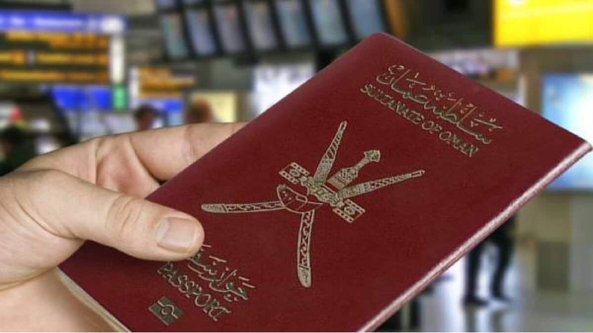 A hand holding an Omani passport at an airport, with departure screens visible in the background.
