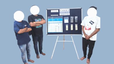 Accused with blurred faces seen standing near a table displaying seized cash and mobile phones in a cricket betting case.