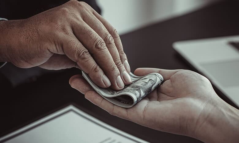 A close-up image of one person handing a rolled bundle of U.S. dollar bills to another person, suggesting a bribe.