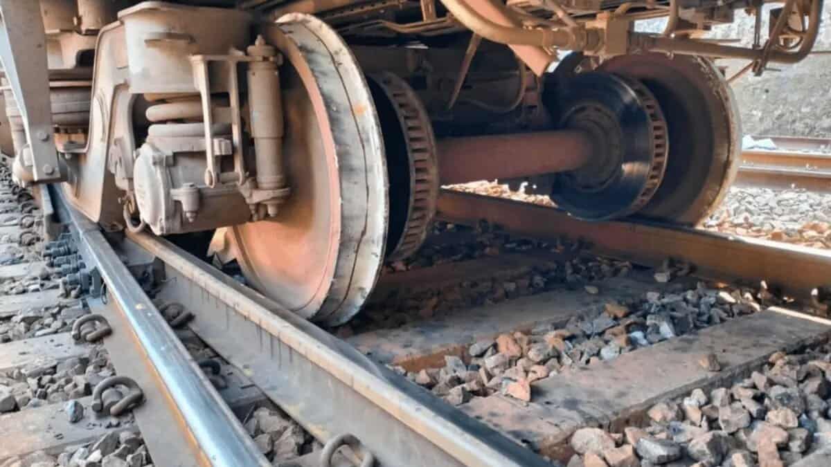 Close-up of a train's undercarriage, showing wheels on tracks with gravel ballast. The mechanical components, including axles and suspension, are visible.