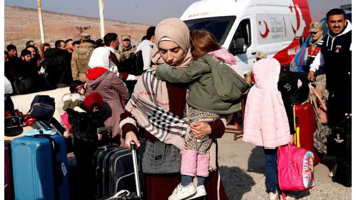 A mother carrying her child walks with luggage amid a crowd of refugees and aid workers near a Turkish Red Crescent van.