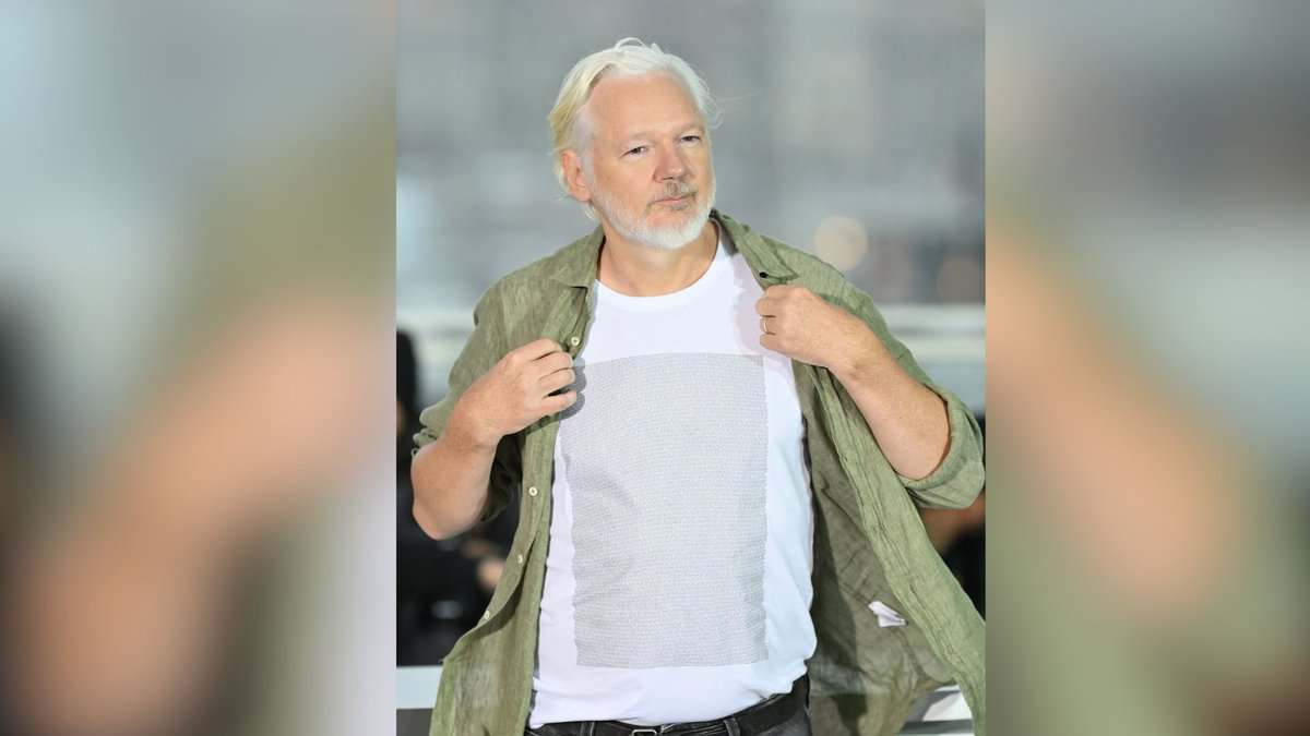 The image displays a picture of WikiLeaks founder Julian Assange at the 78th Cannes Film Festival in France wearing a T-shirt with the names of Palestinian children killed by Israeli forces since 2023.